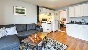 A modern living room with a grey sofa, a wooden coffee table, and a kitchen area with white cabinets and appliances. - Downtown Hallowell Retreat w/ Water Views! (Hallowell)