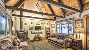 A cozy cabin interior with wooden beams, a fireplace, a large window, and a fireplace mantel with various decorations. - 8 Mi to Heavenly Mtn Resort! Hot Tub Hideaway (South Lake Tahoe)