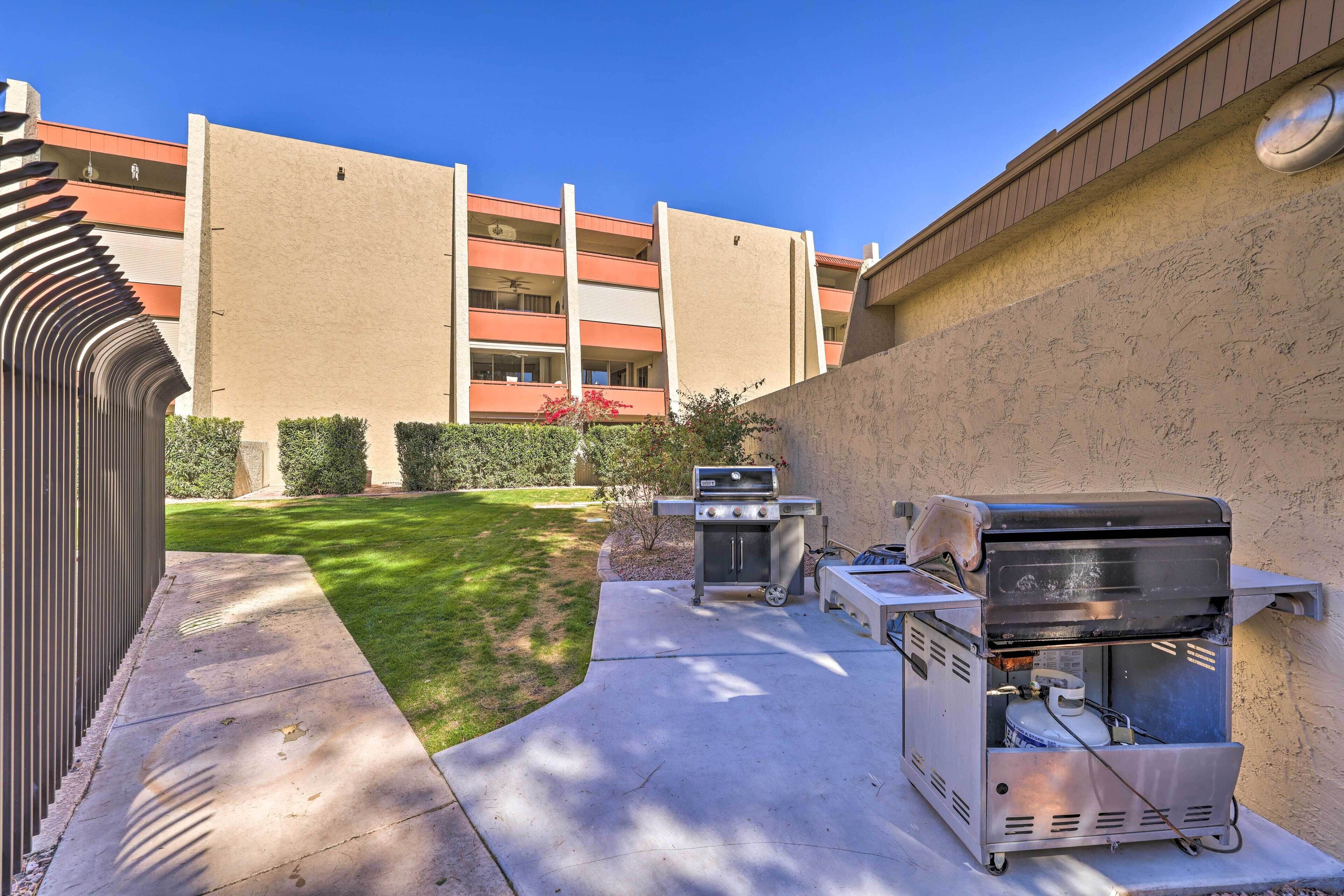 An outdoor patio with a barbecue grill and a grill cart, a concrete pathway, and a building in the background.