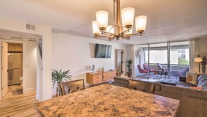 A spacious living room with a large marble dining table, comfortable seating, a chandelier, a TV mounted on the wall, and a view of the outdoors through large windows.