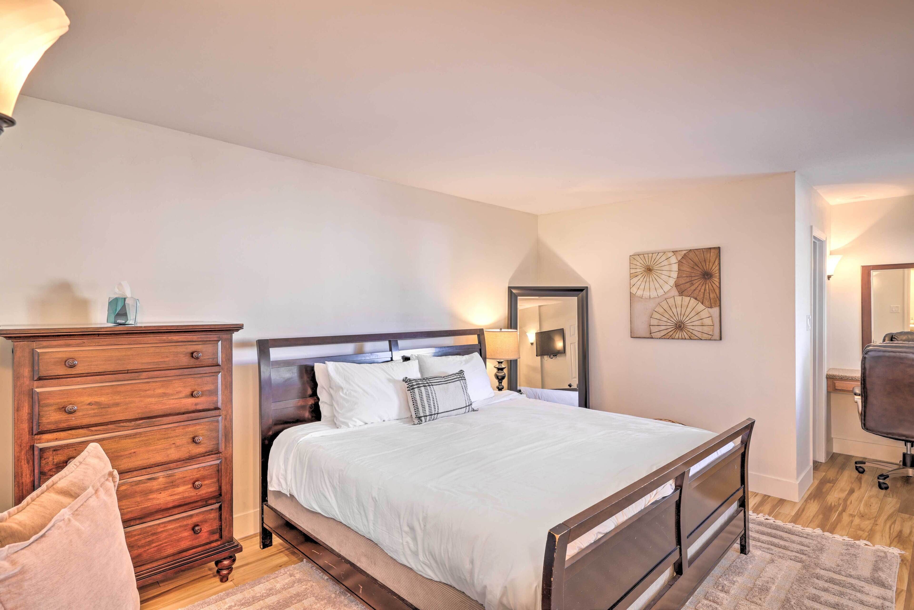 A bedroom with a wooden dresser, a bed with white bedding, a nightstand, and a wall-mounted lamp.