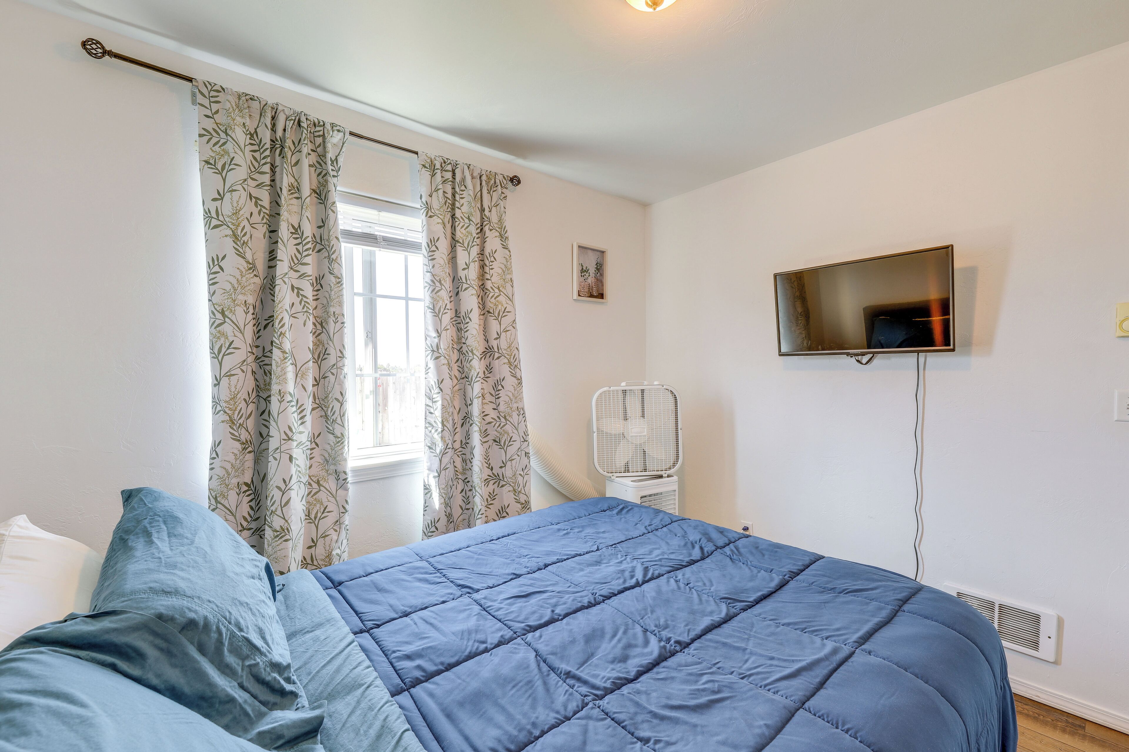 A bedroom with a bed, a TV mounted on the wall, a window with curtains, and a standing fan.