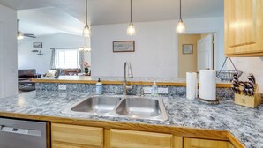 Una cocina con encimeras de granito, lavamanos de acero inoxidable, armarios de madera y luces colgantes, además de una puerta visible que da a otra habitación.