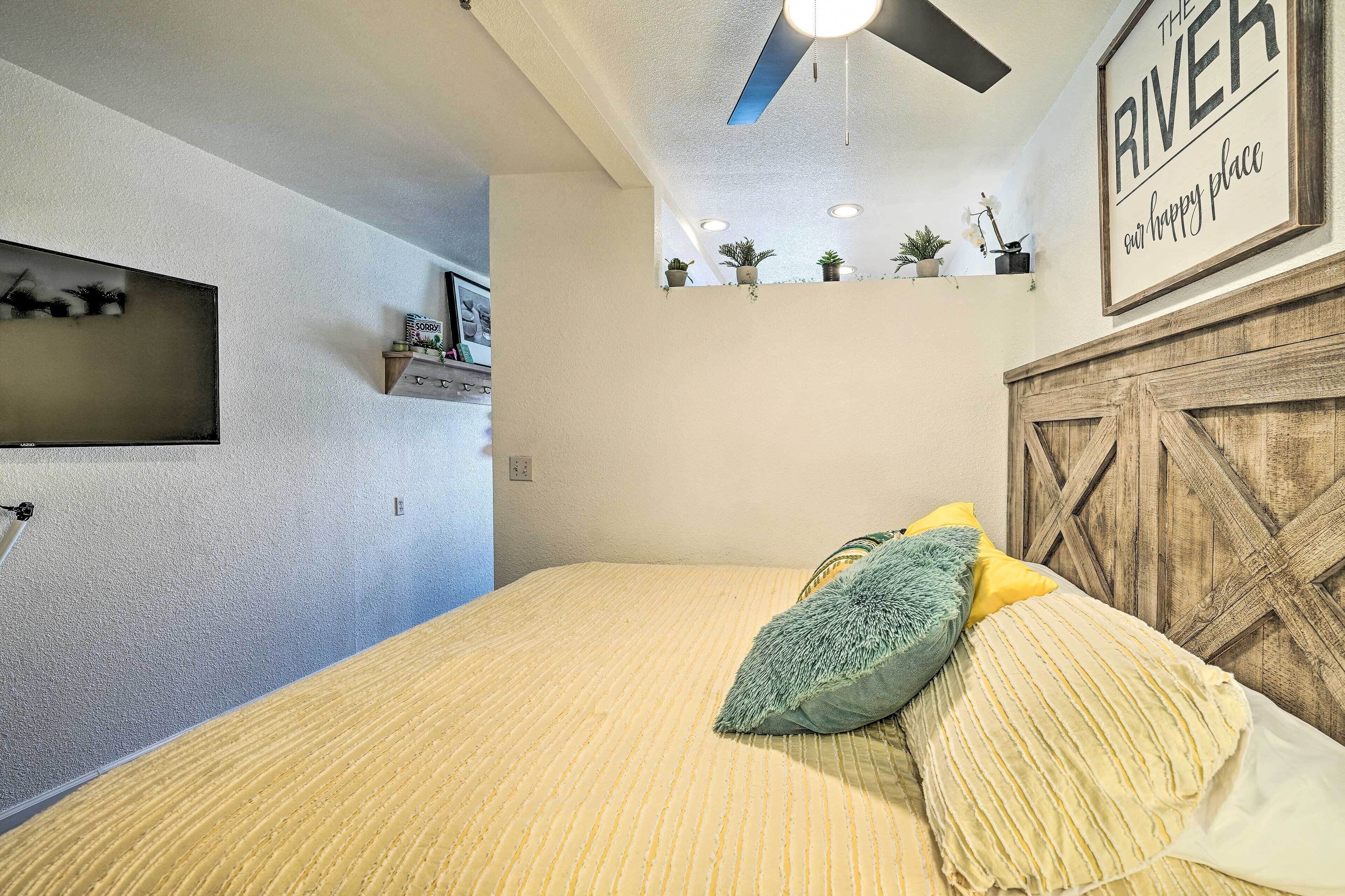 Un dormitorio con cama, almohadas, un estante con plantas y un ventilador de techo, además de un cabecero de madera.