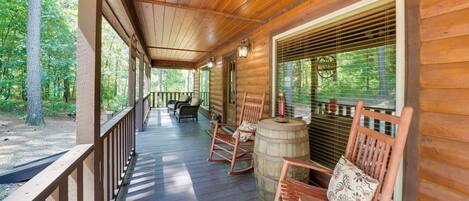 Een houten veranda met schommelstoelen en een vat, omringd door bomen.