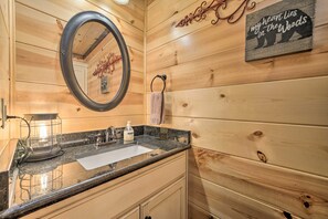 Une salle de bain rustique, avec des murs en bois, un comptoir en granit, un lavabo, un miroir, un porte-serviettes et un panneau décoratif mural.