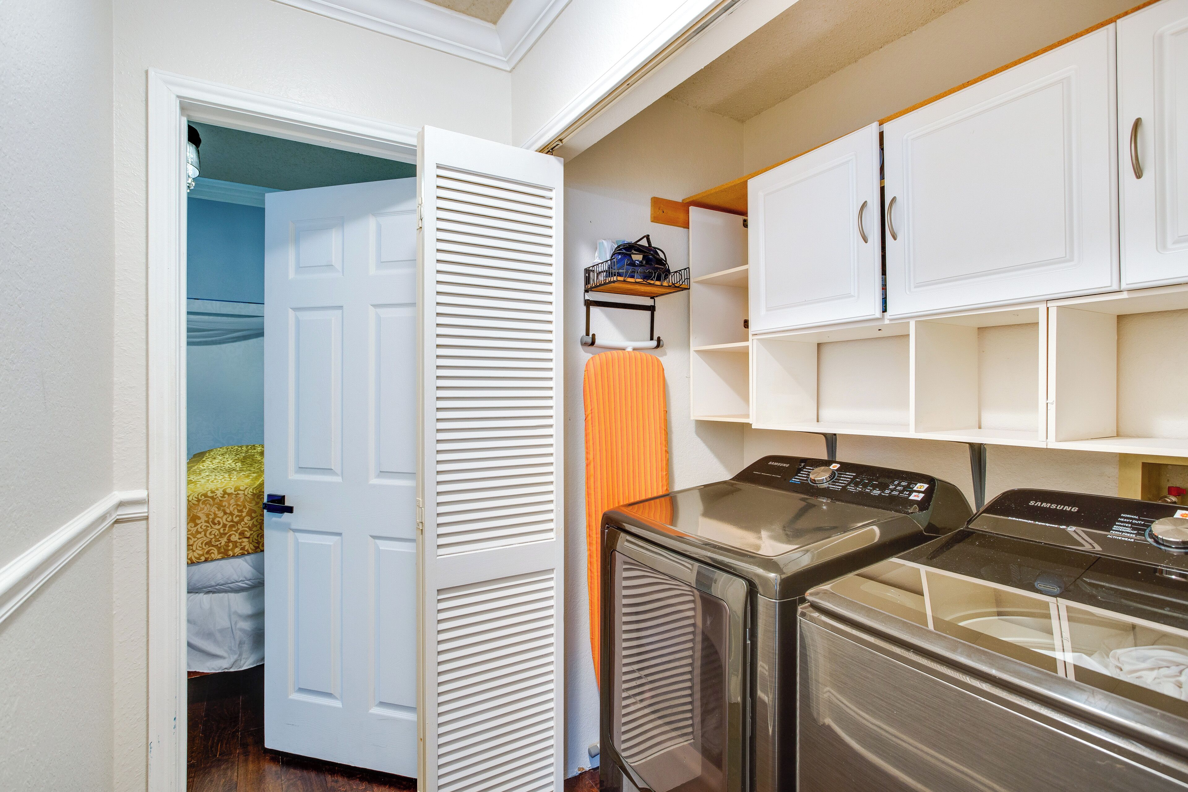 A laundry room with a front-loading washer and dryer, a shelving unit, and a bed in the adjacent room.