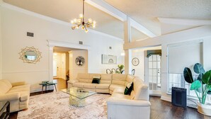 A spacious living room with a large sectional sofa, a glass coffee table, and a chandelier. There are framed pictures on the walls and a potted plant by the window. - Walk to Lake! Rowlett Escape w/ Fire Pit & Views (Rowlett)