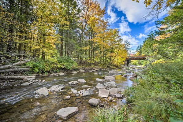 A clear stream with rocks, surrounded by trees with autumn foliage, and a wooden bridge in the background.
