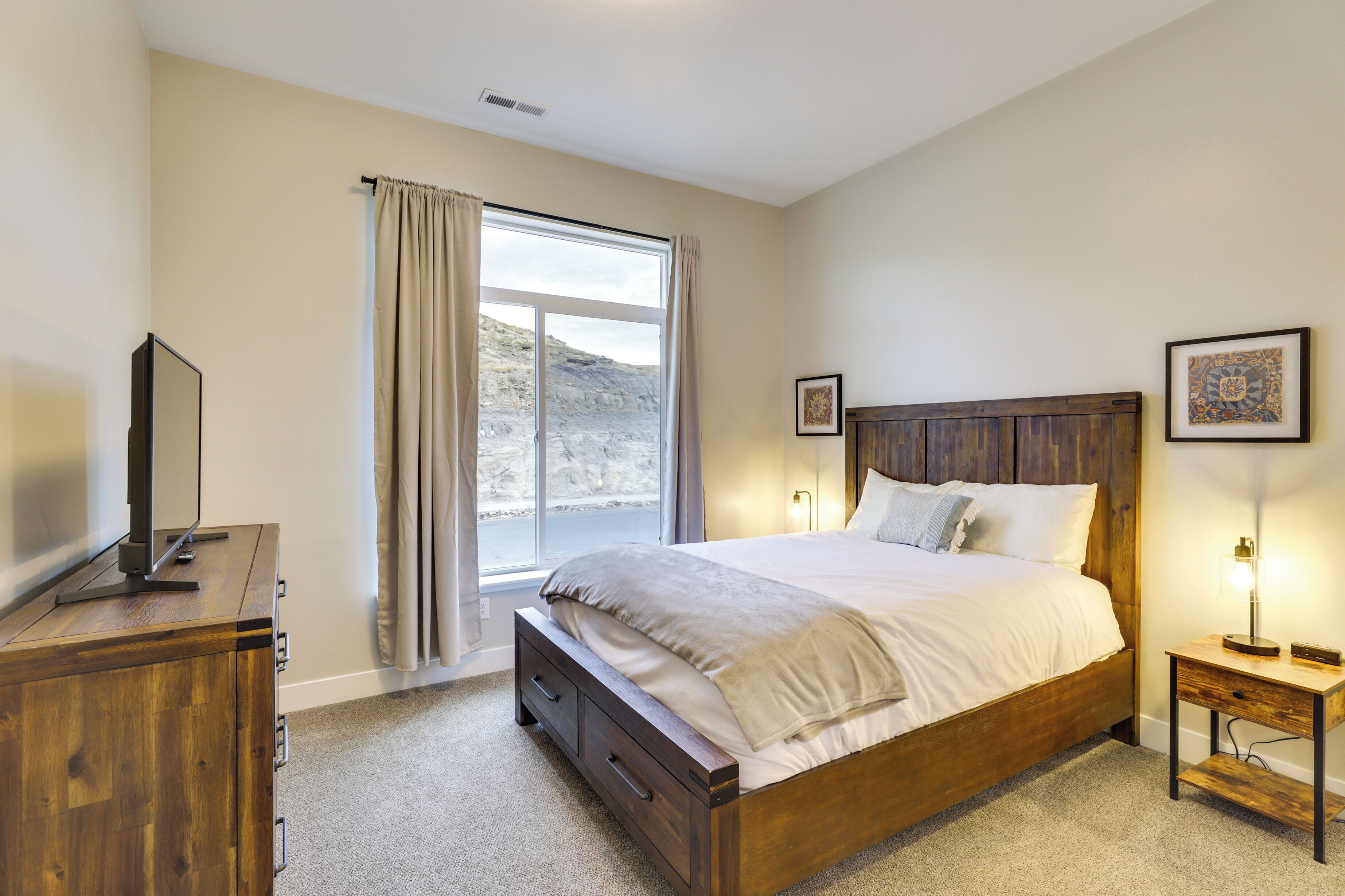 A bedroom with a wooden bed, a nightstand, a dresser, a window with curtains, and a framed picture on the wall.