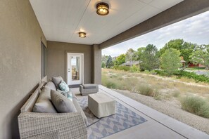 Un espacio de estar al aire libre techado con sofá y reposapiés, una puerta y vistas a un jardín ajardinado y casas.