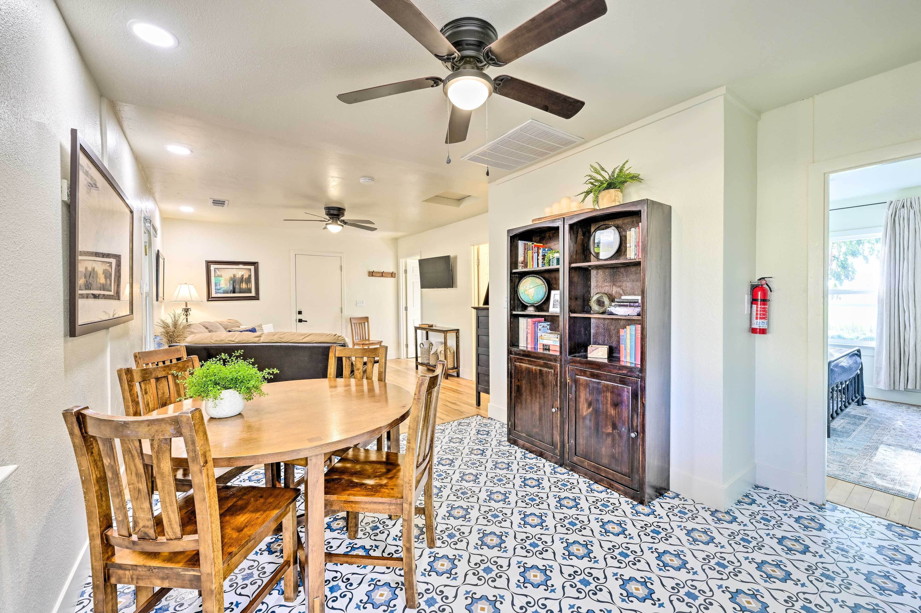 A dining area with a round wooden table, chairs, and a ceiling fan. There is a bookshelf with books and decorative items. The floor has a patterned tile design.