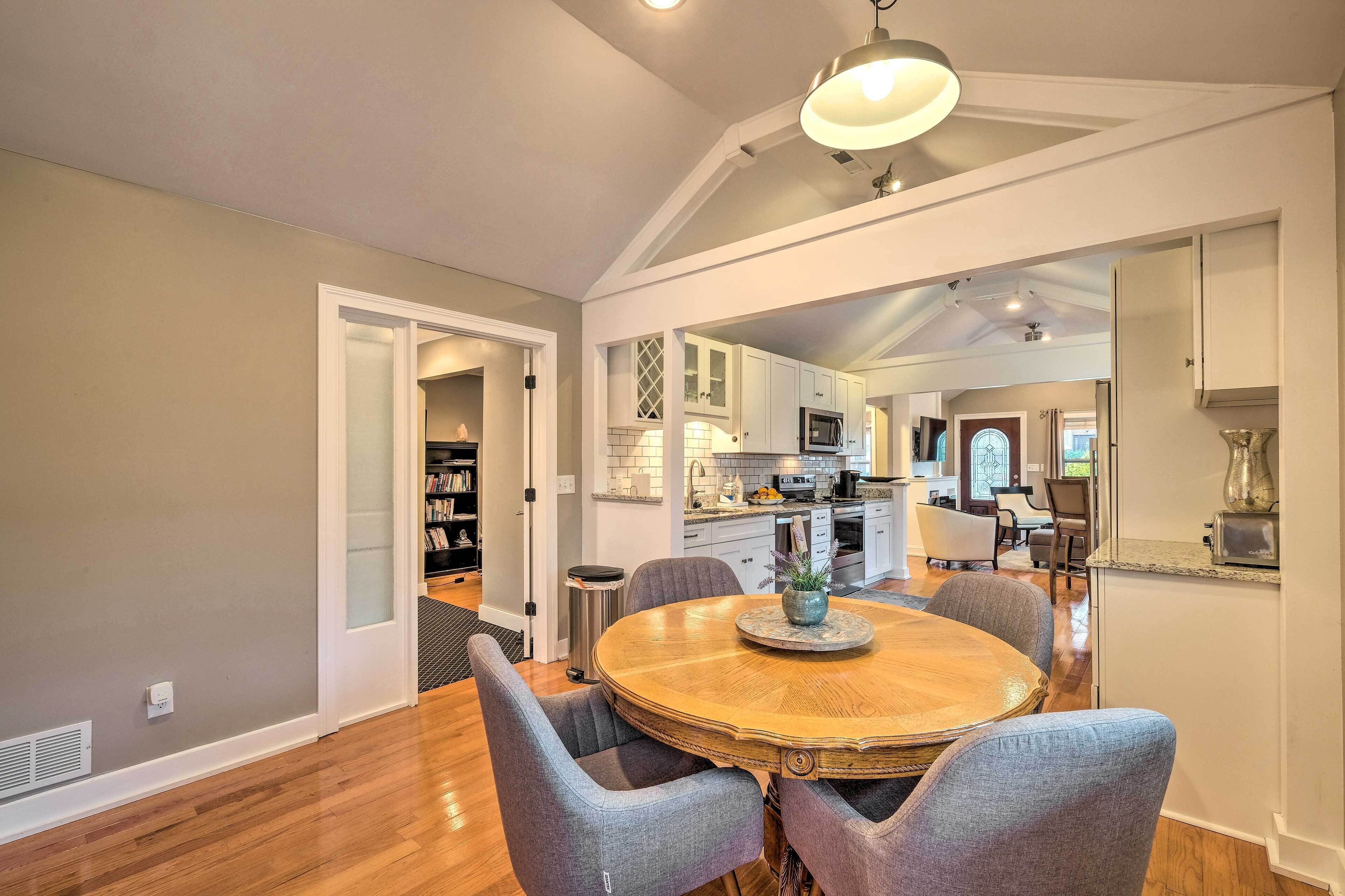A dining area with a round wooden table and grey upholstered chairs, a kitchen in the background, and a ceiling with exposed beams and recessed lighting.