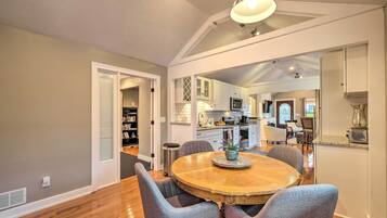 A dining area with a round wooden table and grey upholstered chairs, a kitchen in the background, and a ceiling with exposed beams and recessed lighting.