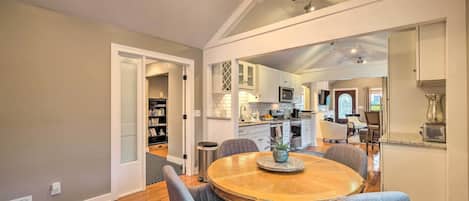 A dining area with a round wooden table and grey upholstered chairs, a kitchen in the background, and a ceiling with exposed beams and recessed lighting.