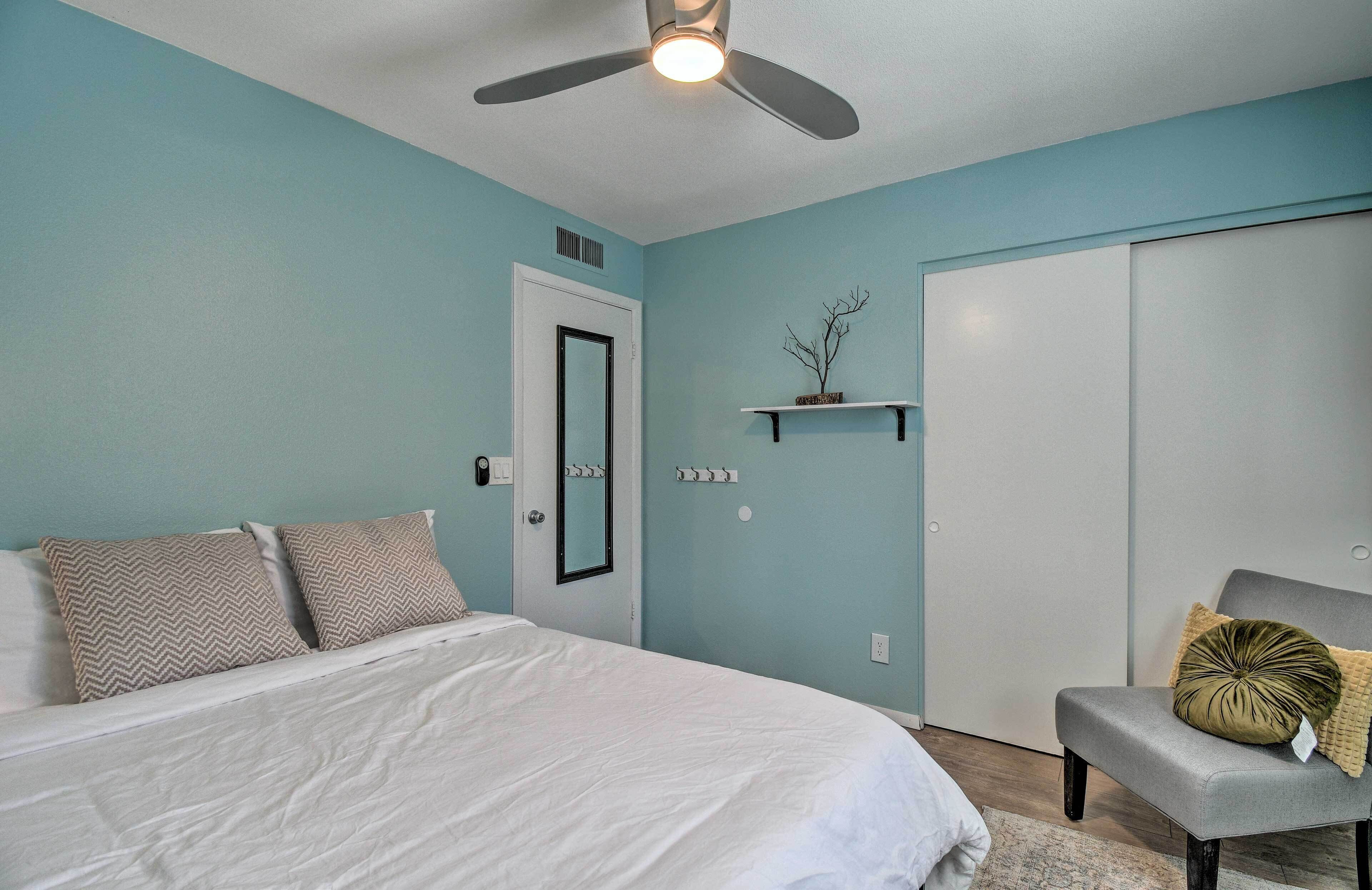 Un dormitorio con cama, una silla, ventilador de techo, una puerta y una estantería con una planta.