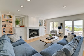 A modern living room with a fireplace, blue sofas, and a view of the outdoors. - Finger Lakes Vacation Rental w/ Hot Tub & Pool (Naples)