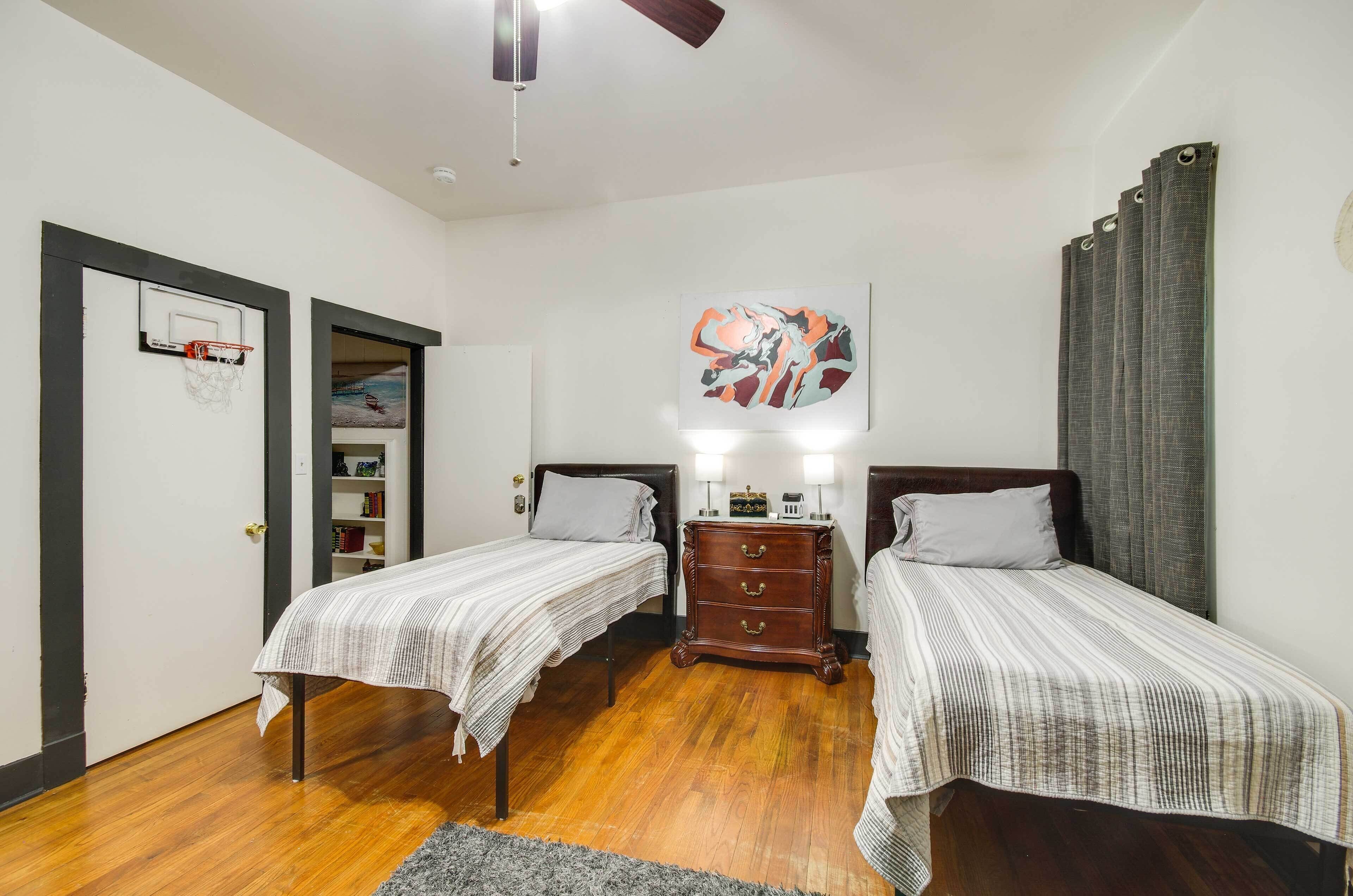 A bedroom with two single beds, a wooden dresser, a ceiling fan, a basketball hoop, and a large abstract painting.