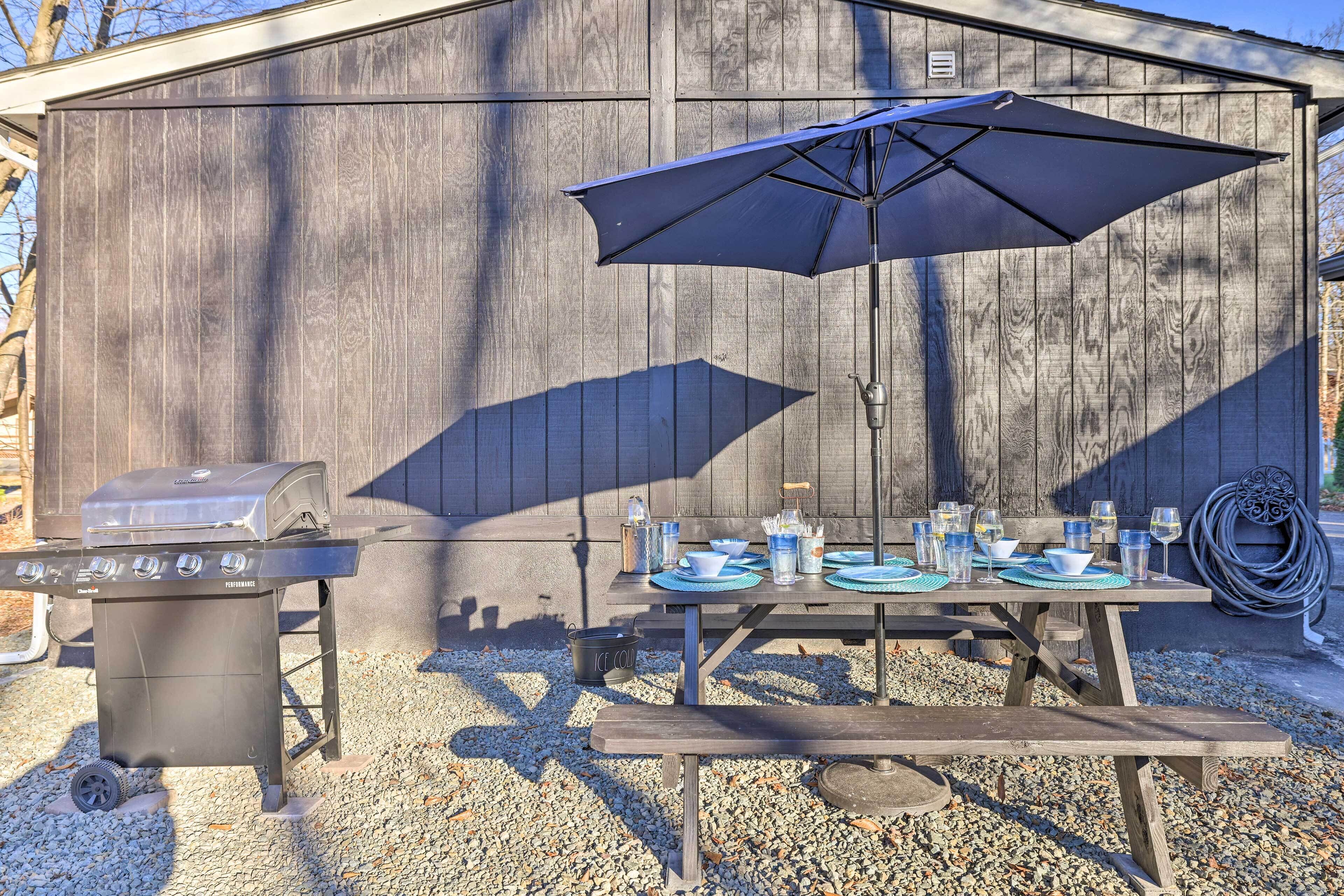An outdoor barbecue setup with a grill, picnic table, umbrella, and various dining items.