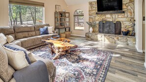 A spacious living room with a large sectional sofa, a wooden coffee table, and a stone fireplace with a flat-screen TV mounted above it. - Deck + Game Room: Lakeside 'blue Jay Chalet' Cabin (Lakeside)