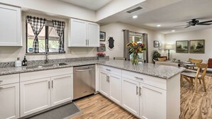 A modern kitchen with white cabinets, granite countertops, and a dishwasher. There is a dining area with a table and chairs, and a ceiling fan. - Sunny Phoenix Home w/ Pool + Backyard Oasis! (Phoenix)