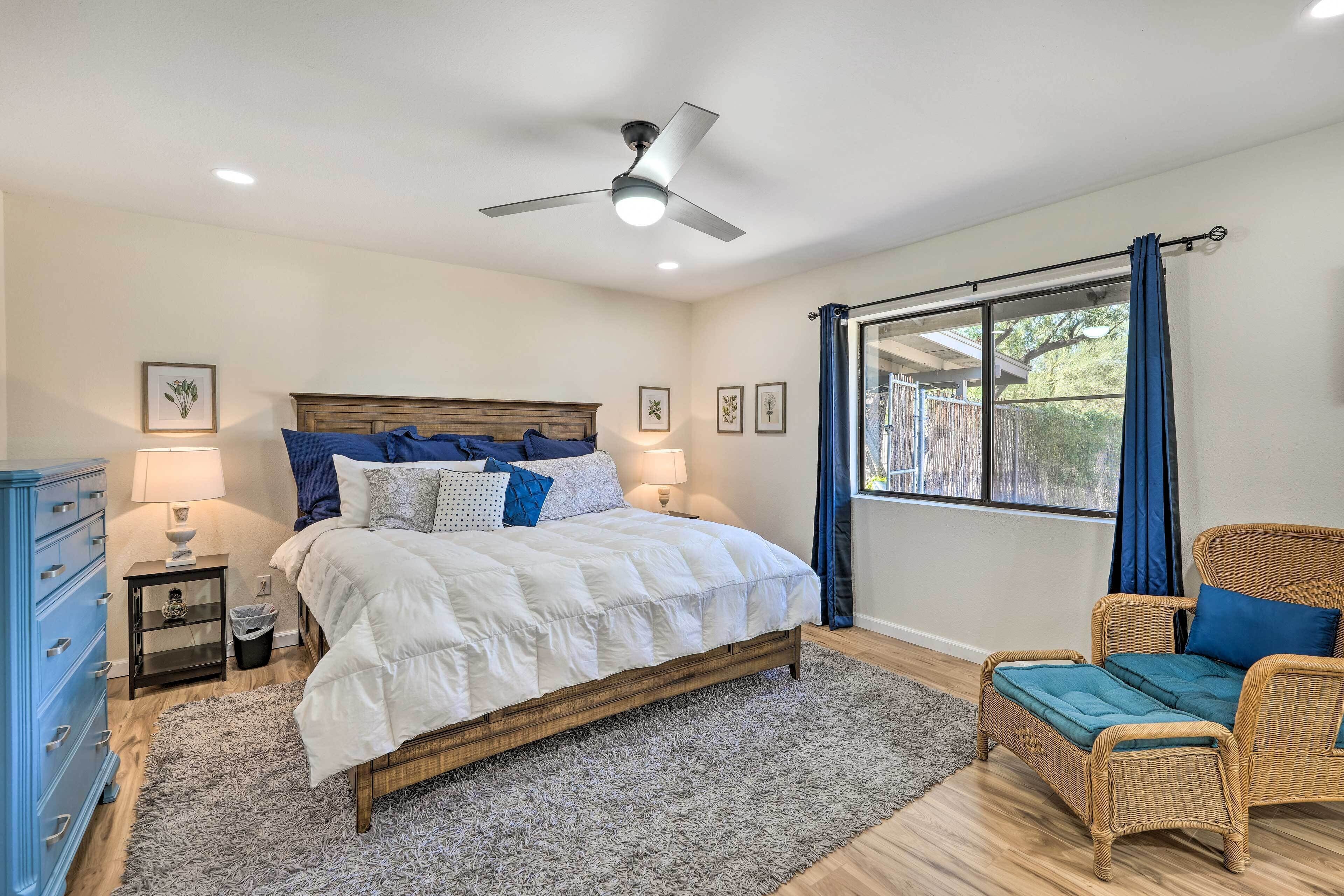 Une chambre à coucher comprenant un lit, une chaise, un ventilateur de plafond, une fenêtre avec des rideaux et une table de chevet avec une lampe.