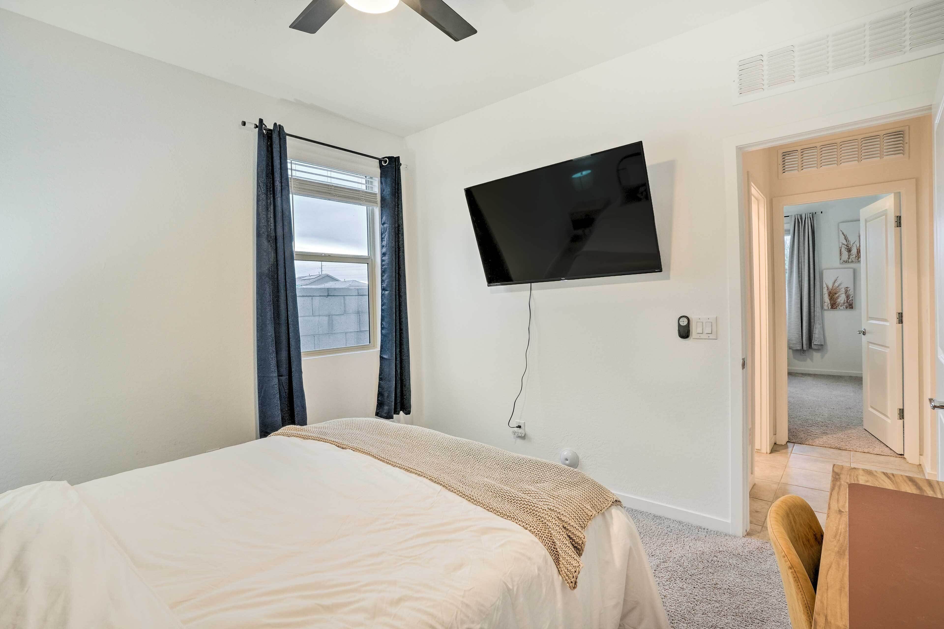 Un dormitorio con una cama, un televisor montado en la pared, una ventana con cortinas y una puerta que da a otra habitación.