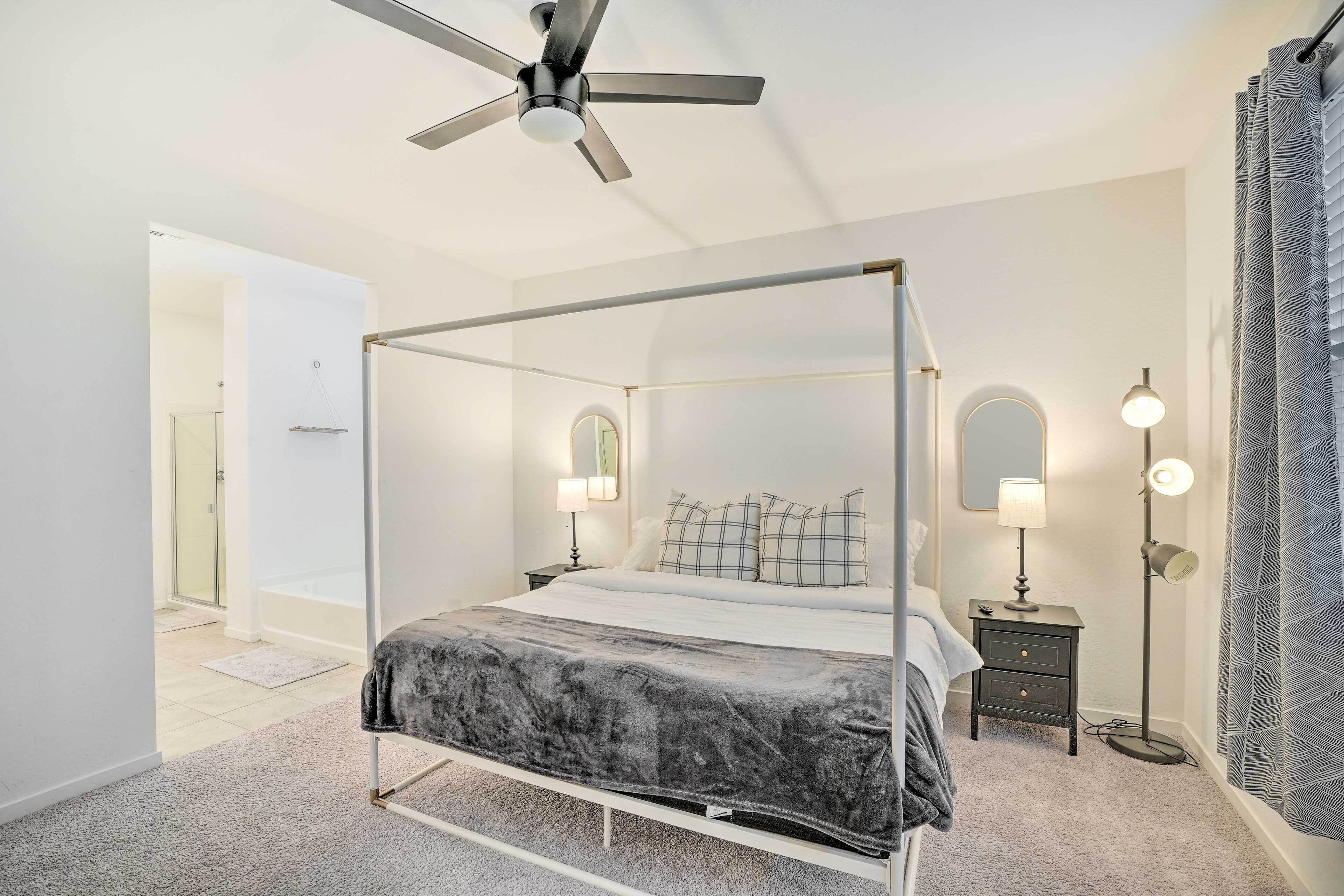 Una cama con dosel y un edredón tipo mármol, una lámpara de pie y un ventilador de techo en un dormitorio con vestidor y un baño visible a través de una puerta abierta.