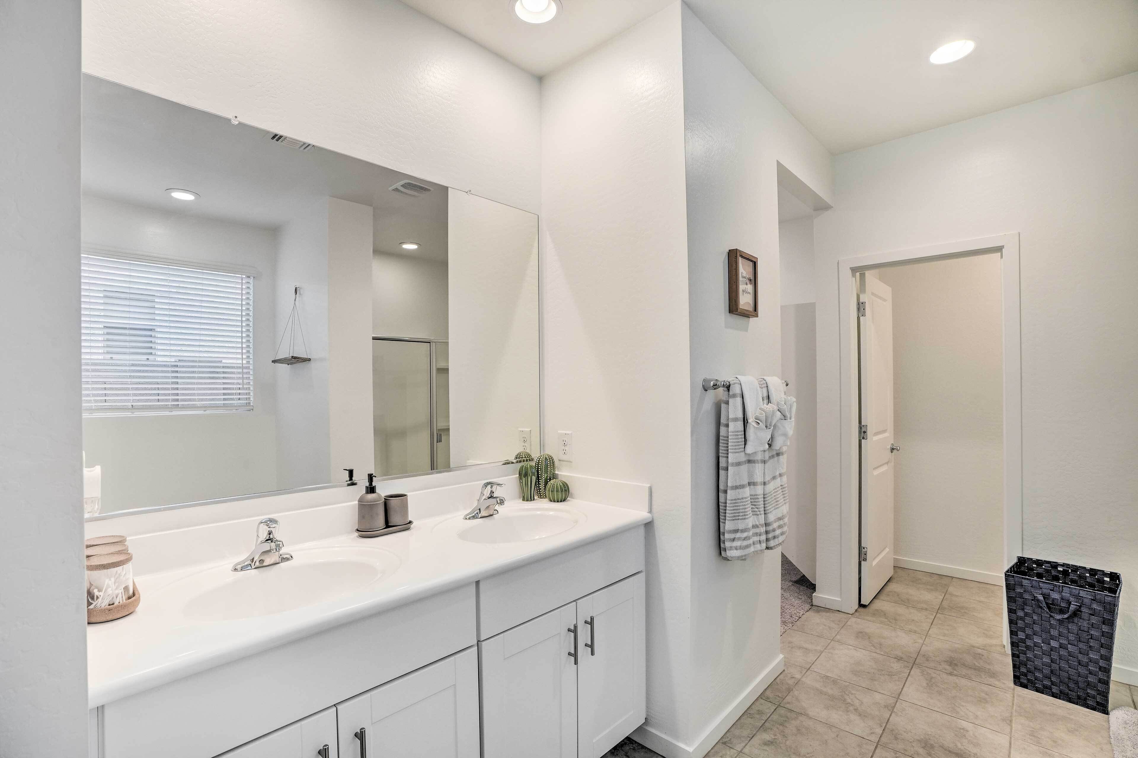 A modern bathroom with a large mirror, white vanity, and a sink. There is a towel rack with a towel hanging, and a trash bin is visible.