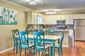 A kitchen with white cabinets, a stainless steel refrigerator, a microwave, and a stove. There is a dining table with chairs and a trash can.