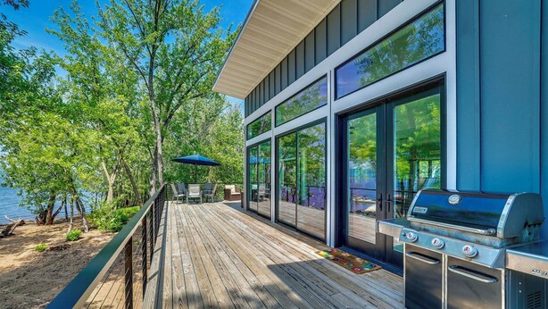 Una terraza con una parrilla, mobiliario exterior y grandes ventanales con vistas a un cuerpo de agua.