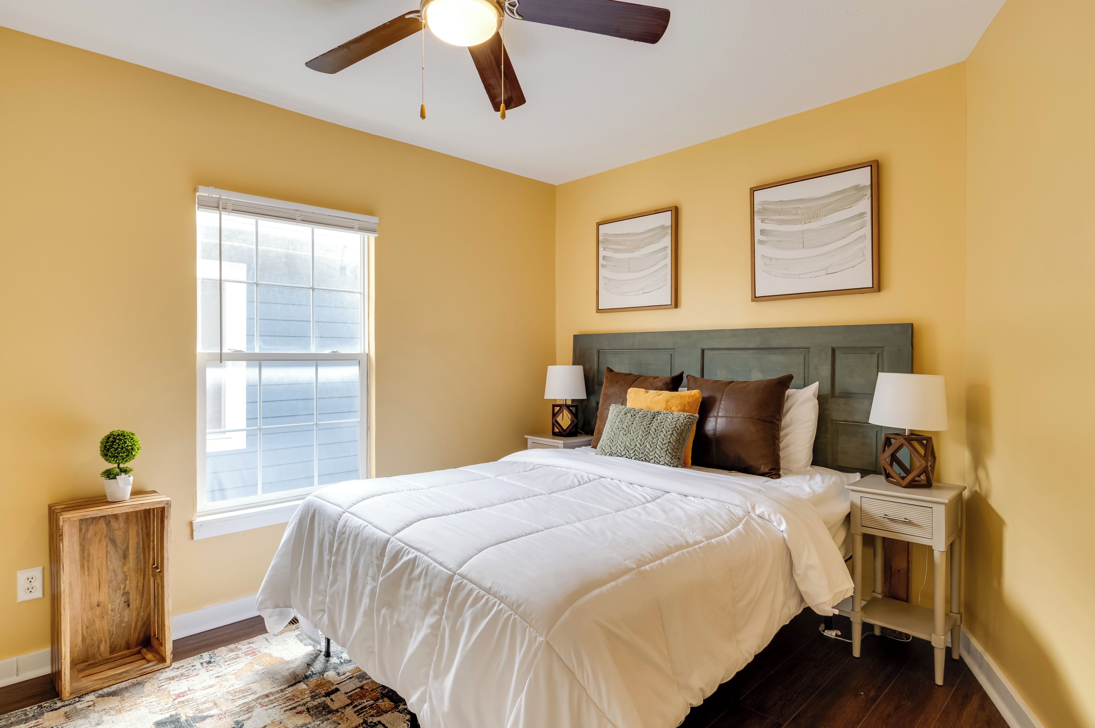 Une chambre à coucher comprenant un lit, deux tables de chevet, un ventilateur de plafond, une fenêtre et un petit meuble.