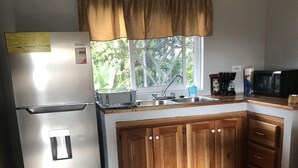 A compact kitchen with a refrigerator, wooden cabinets, a sink, and a window with a view of greenery. - Seacret Studios - Cozy Jamaican Retreat Apts by RedAwning (Negril)