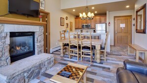 A living room with a stone fireplace, a television mounted on the wall, a dining area with wooden chairs and a table, and a kitchen in the background. - Cozy Avon Condo Near Beaver Creek Resort! (Avon)