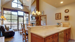A spacious kitchen with a large island, wooden cabinets, and a built-in dishwasher. There is a dining area with a black leather couch and wooden chairs. A large window provides a view of the outdoors. - Cozy Avon Condo Near Beaver Creek Resort! (Avon)