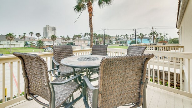 A balcony with wicker chairs and a table, overlooking a residential area with palm trees and buildings.