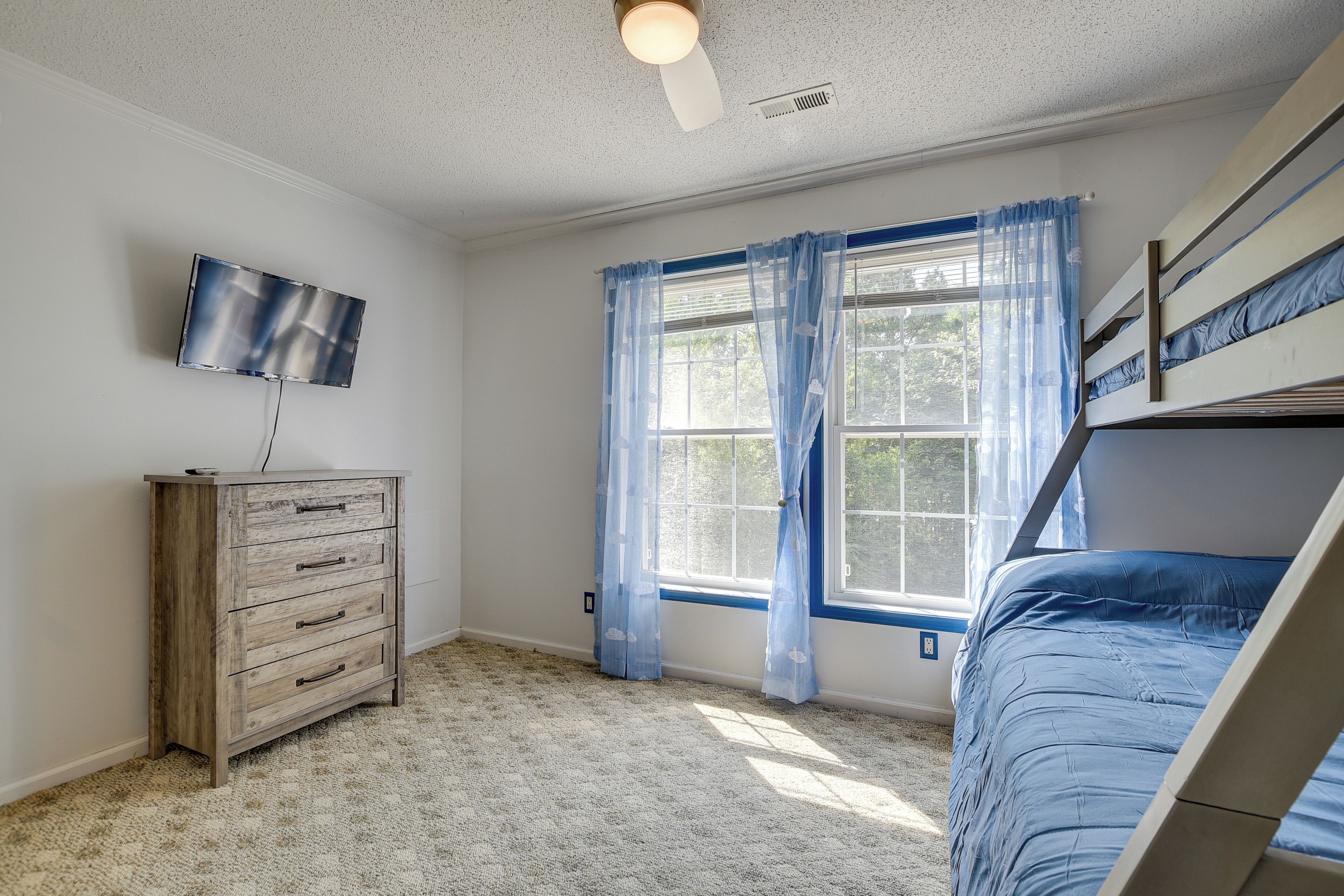 Une chambre avec un lit superposé, une commode en bois, un téléviseur à écran plat et une fenêtre avec des rideaux bleus.