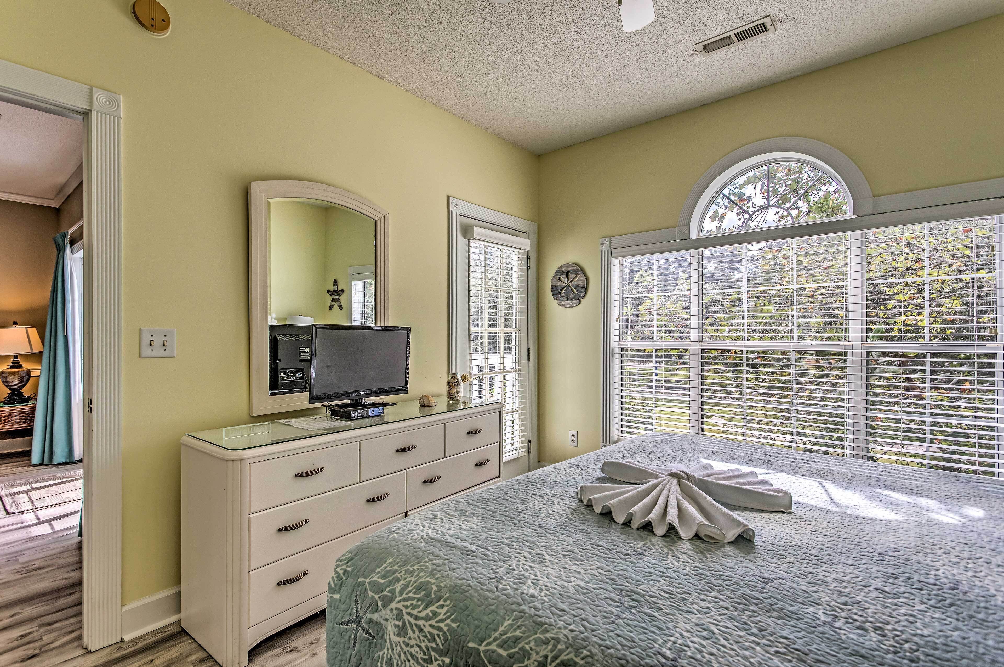 A bedroom with a bed, dresser, television, and large windows with blinds.