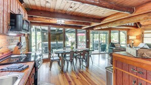 A spacious kitchen with wooden cabinets, a dining area with chairs, and a large window providing a view of the outdoors. - Furnished Deck + Lake Access: Fairland Log Cabin (Fairland)