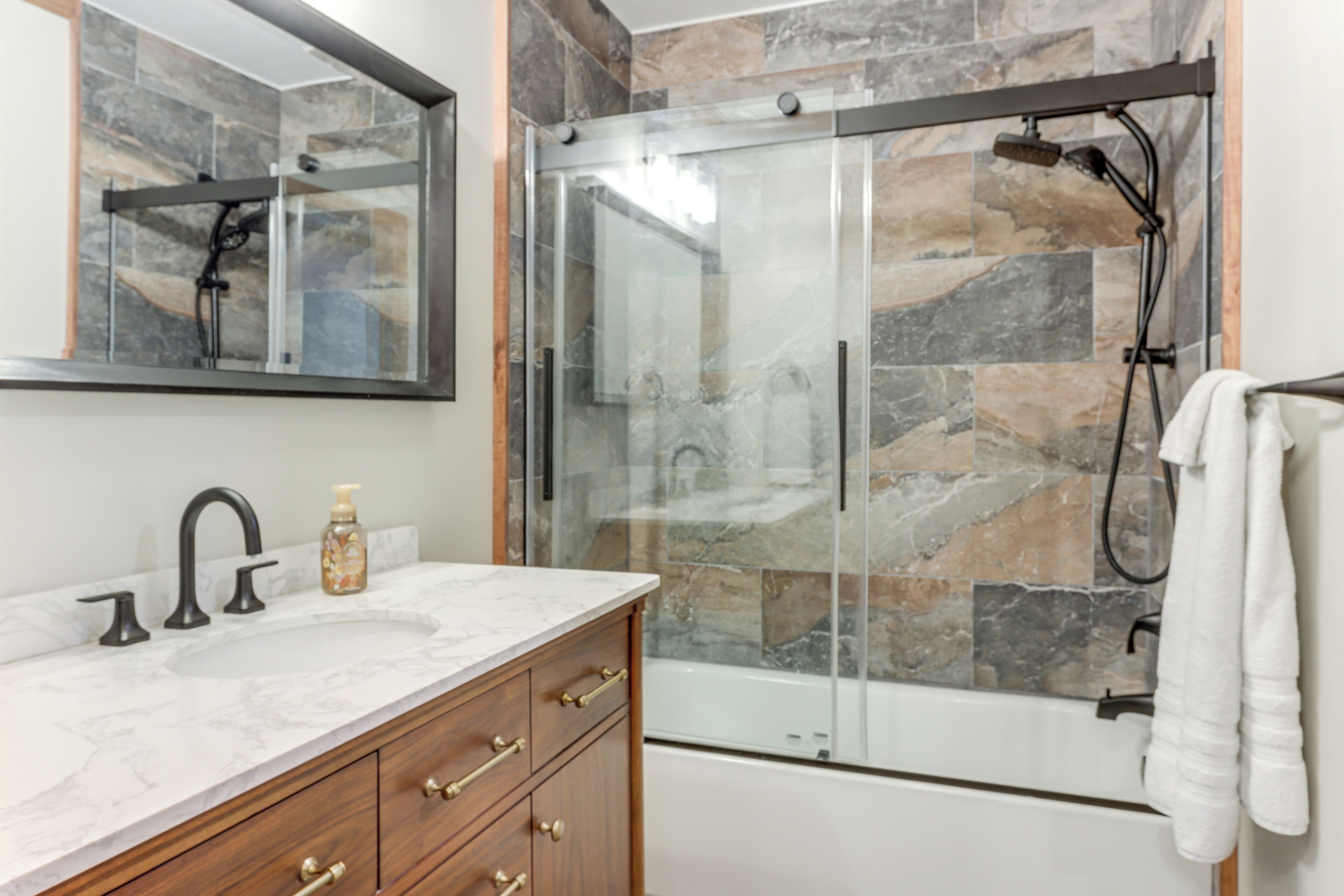 A bathroom with a marble sink, wooden vanity, glass shower enclosure, and a stone-tiled shower wall.