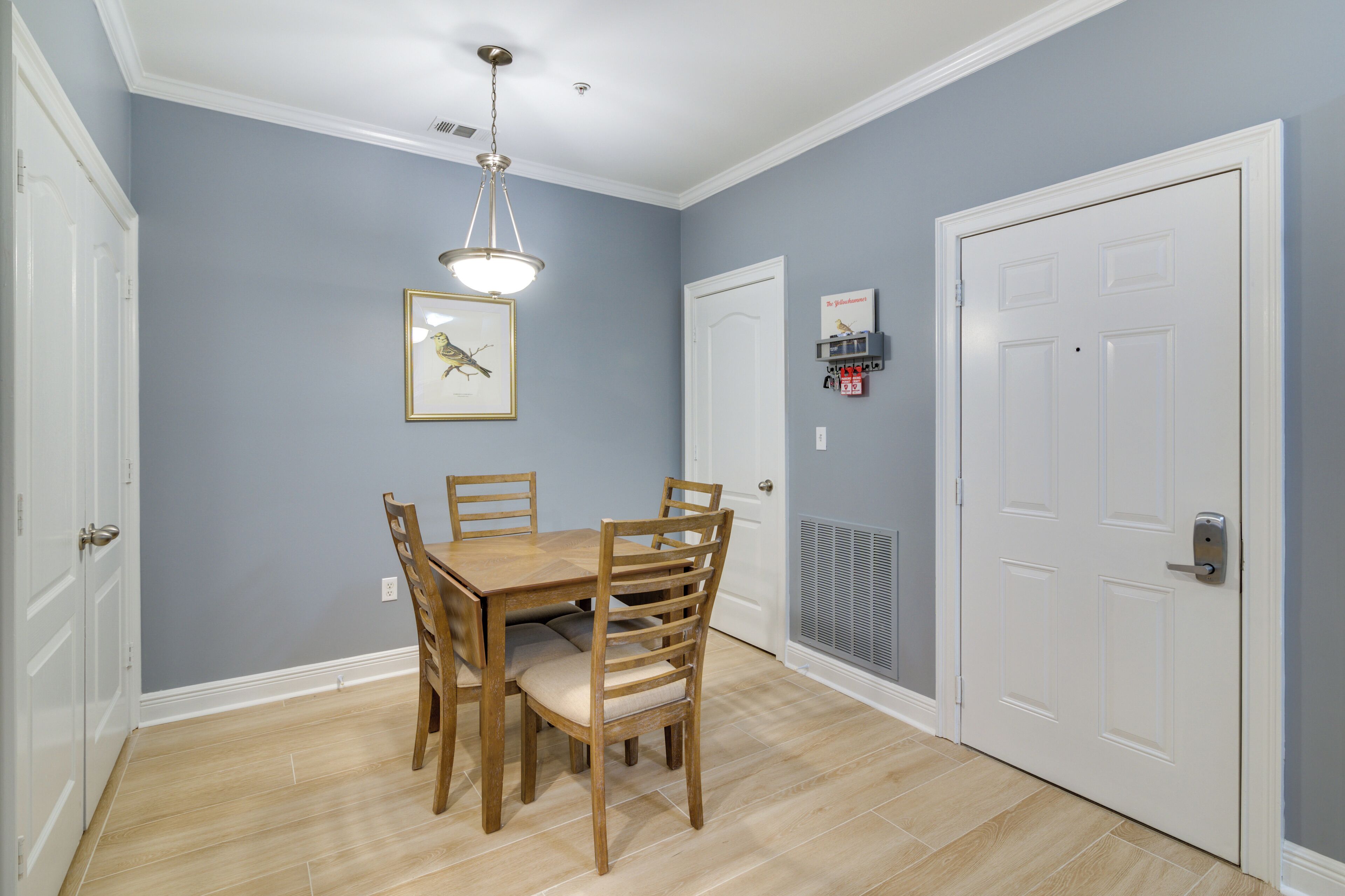 A dining area with a wooden table and chairs, a framed picture on the wall, and a white door with a vent.