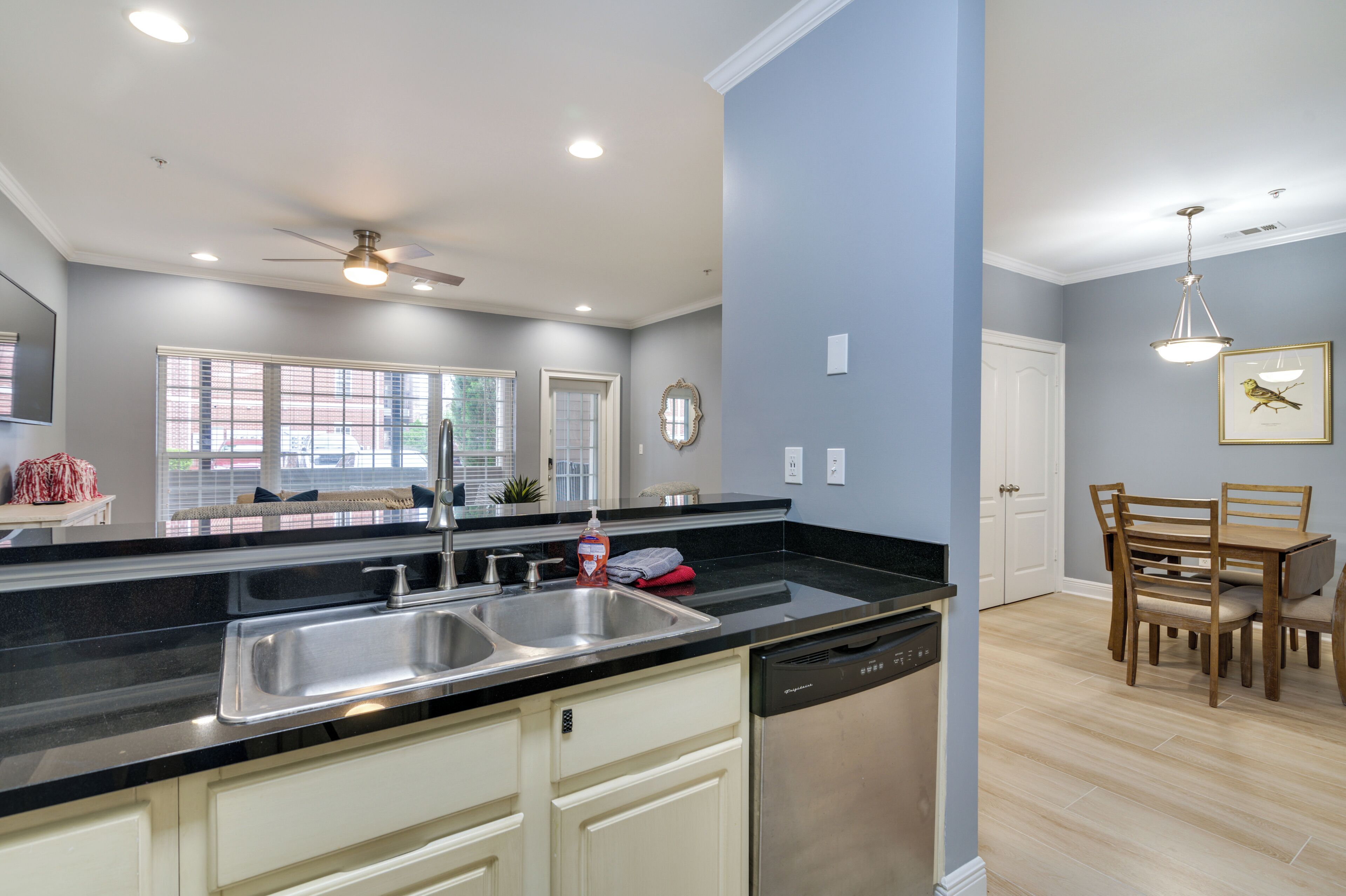A modern kitchen with a black countertop, stainless steel sink, and dishwasher. There is a dining area with a wooden table and chairs.