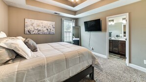 A bedroom with a bed, a ceiling fan, a painting, a TV, and a bathroom visible through an open door.