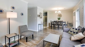 A modern living room with a sofa, coffee table, and a side table with a lamp. There is a dining area with a table and chairs, and a kitchen in the background. - North Charleston Vacation Rental Near Cooper River (North Charleston)