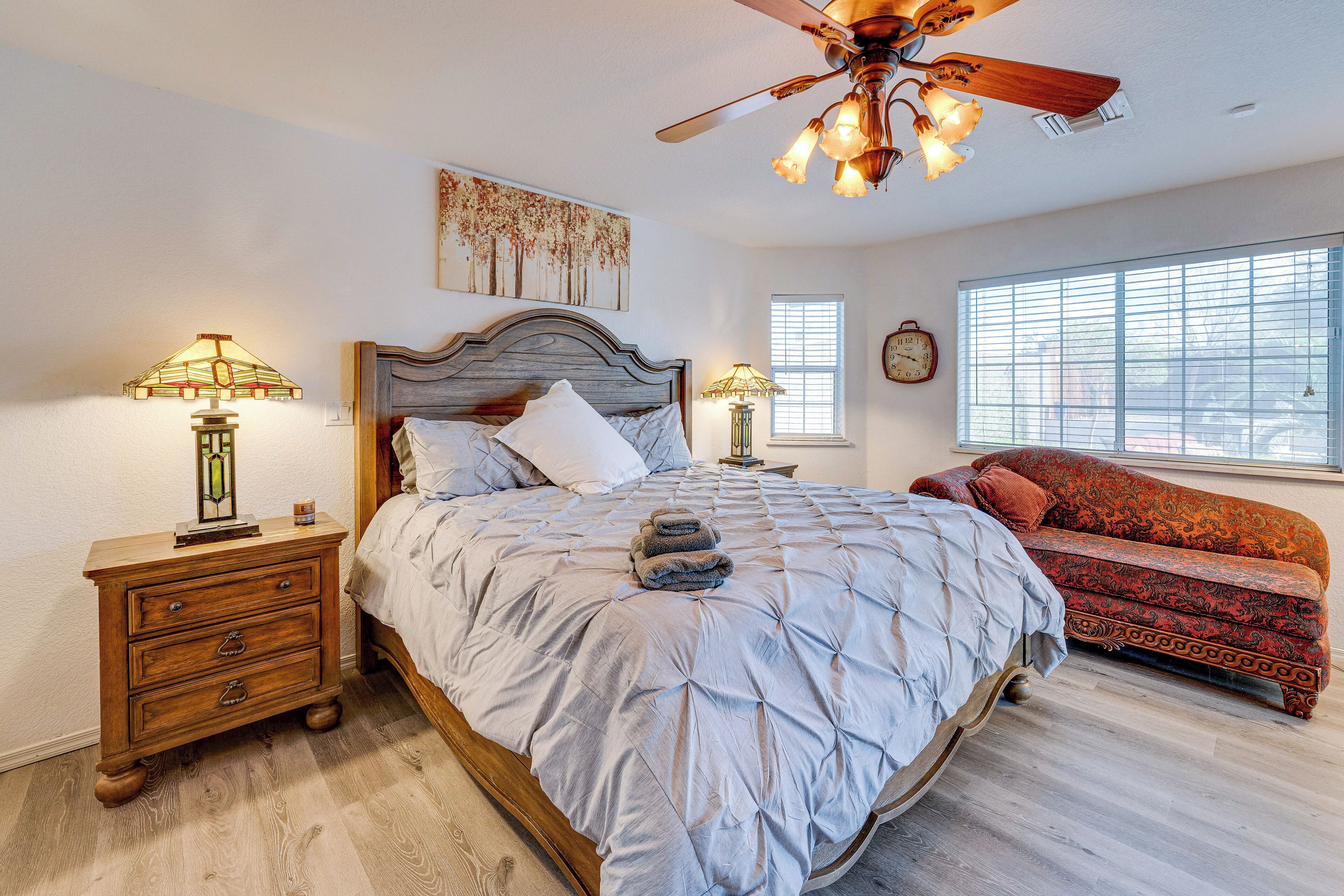 A bedroom with a large bed, a wooden nightstand, a floor lamp, a ceiling fan, and a window with blinds.