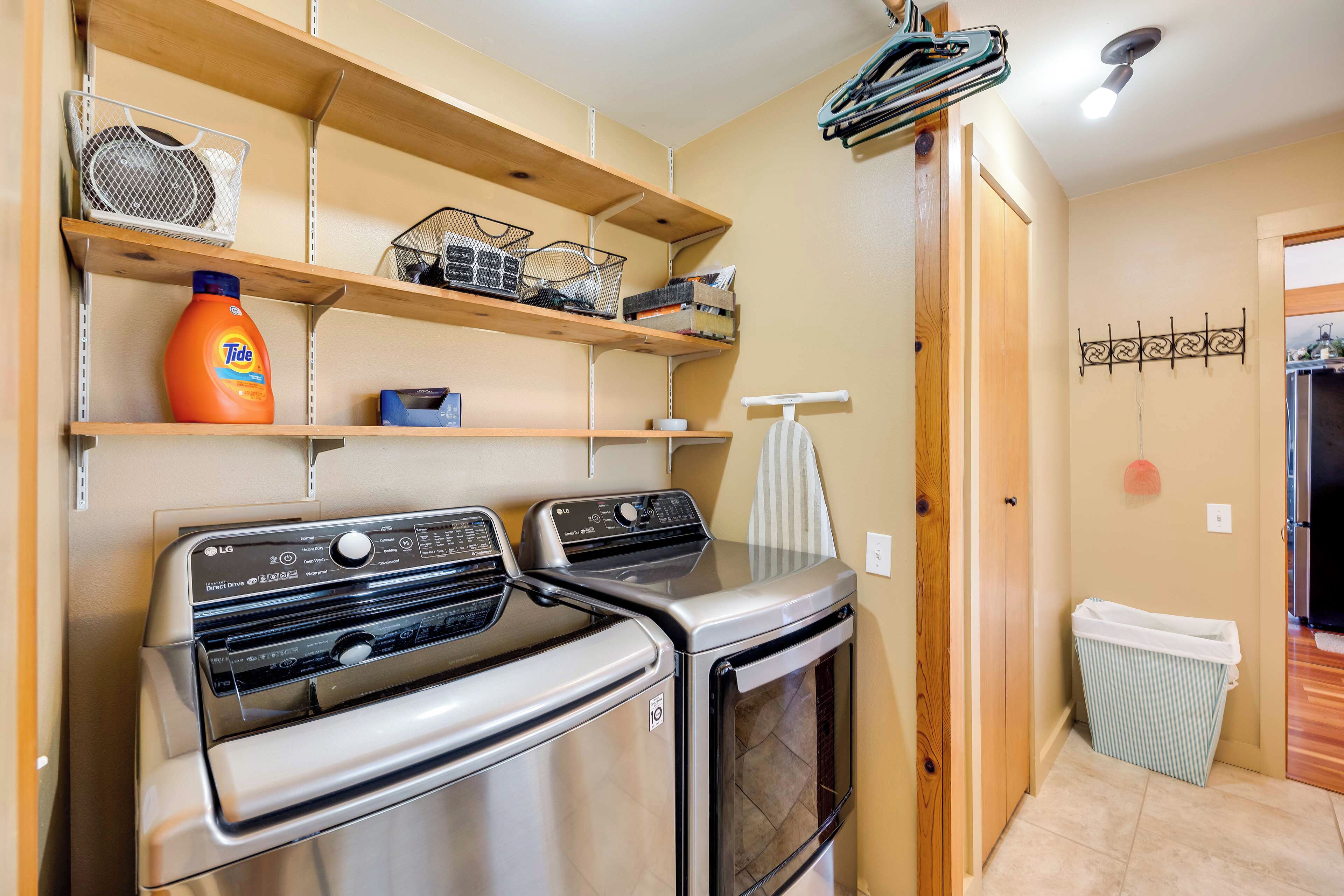 A laundry area with two stainless steel dryers, wooden shelves above holding baskets and a Tide detergent bottle, and a trash bin to the right.