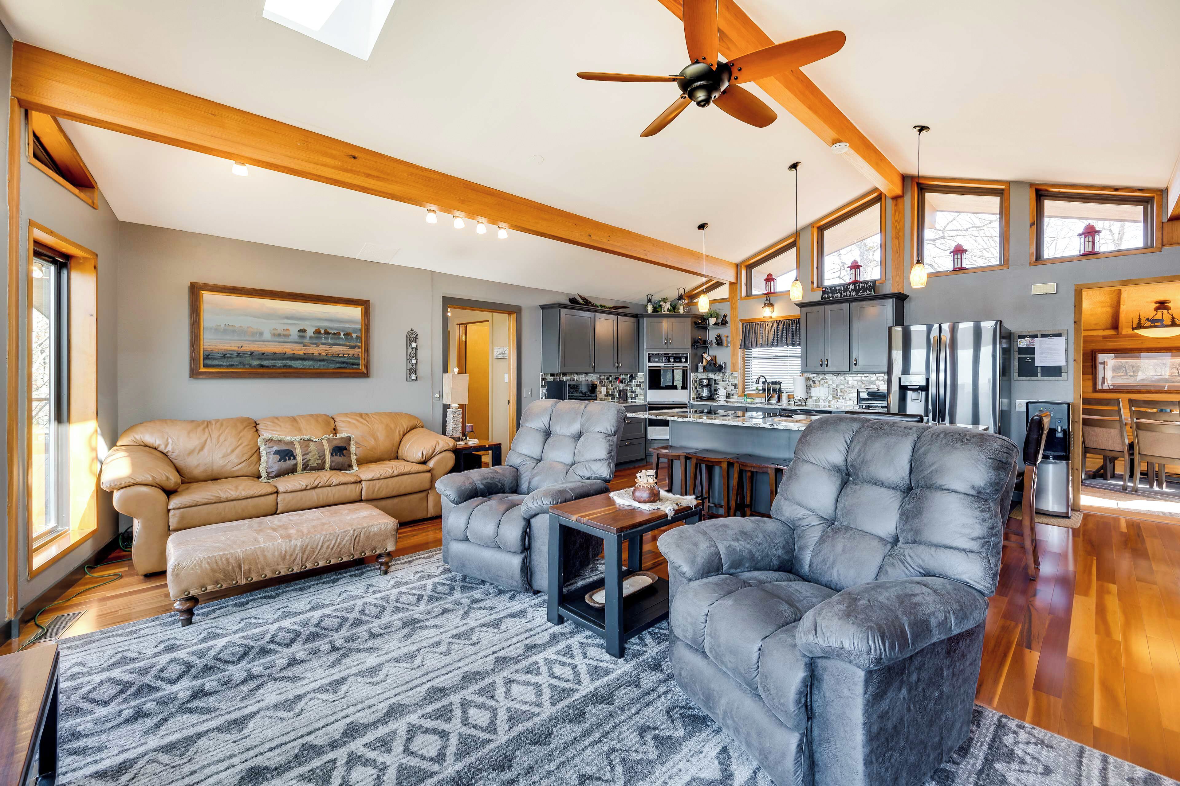 A spacious living room with a sofa, armchairs, and a coffee table. There is a ceiling fan, wooden beams, and a kitchen area with appliances.