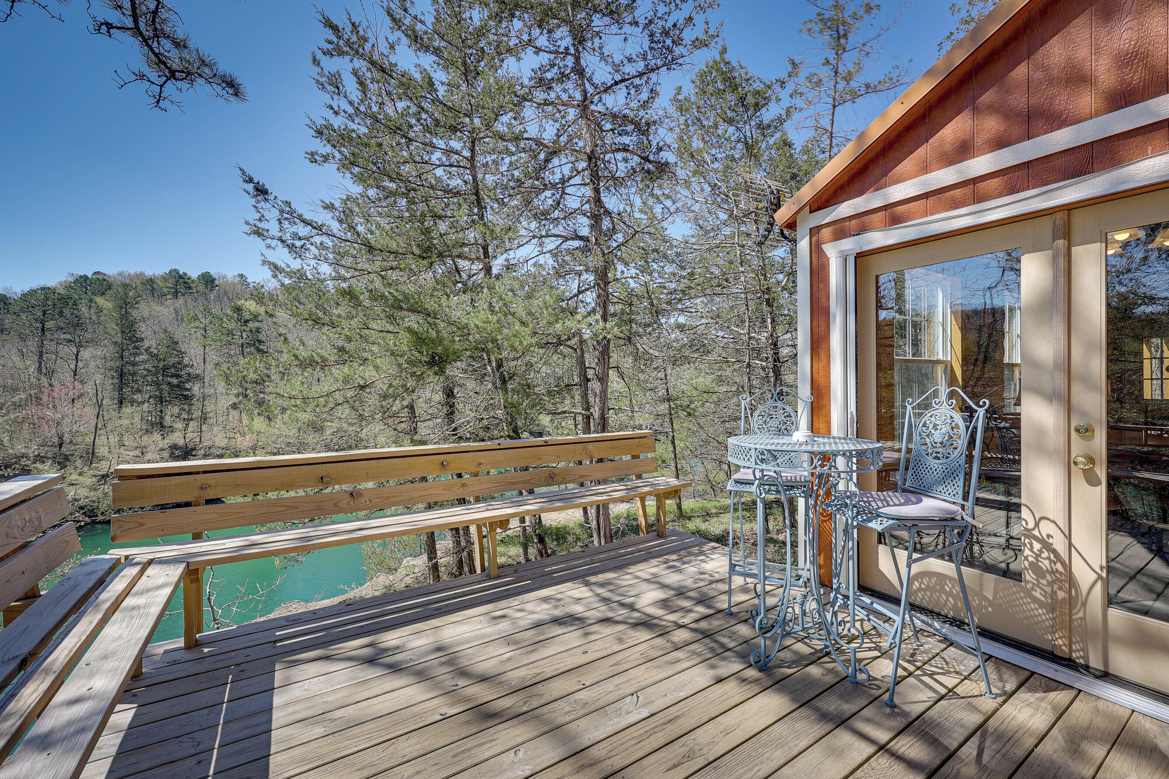 Eine Holzterrasse mit einer Bank, einem Metalltisch und Stühlen mit Blick auf ein Schwimmbecken und einen Wald.