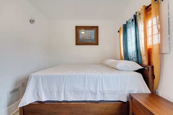 A neatly made bed with a white bedspread and pillows, a wooden headboard, a framed picture on the wall, and a window with curtains. - Buffalo Vacation Rental ~ 20 Mi to Niagara Falls! (Buffalo)