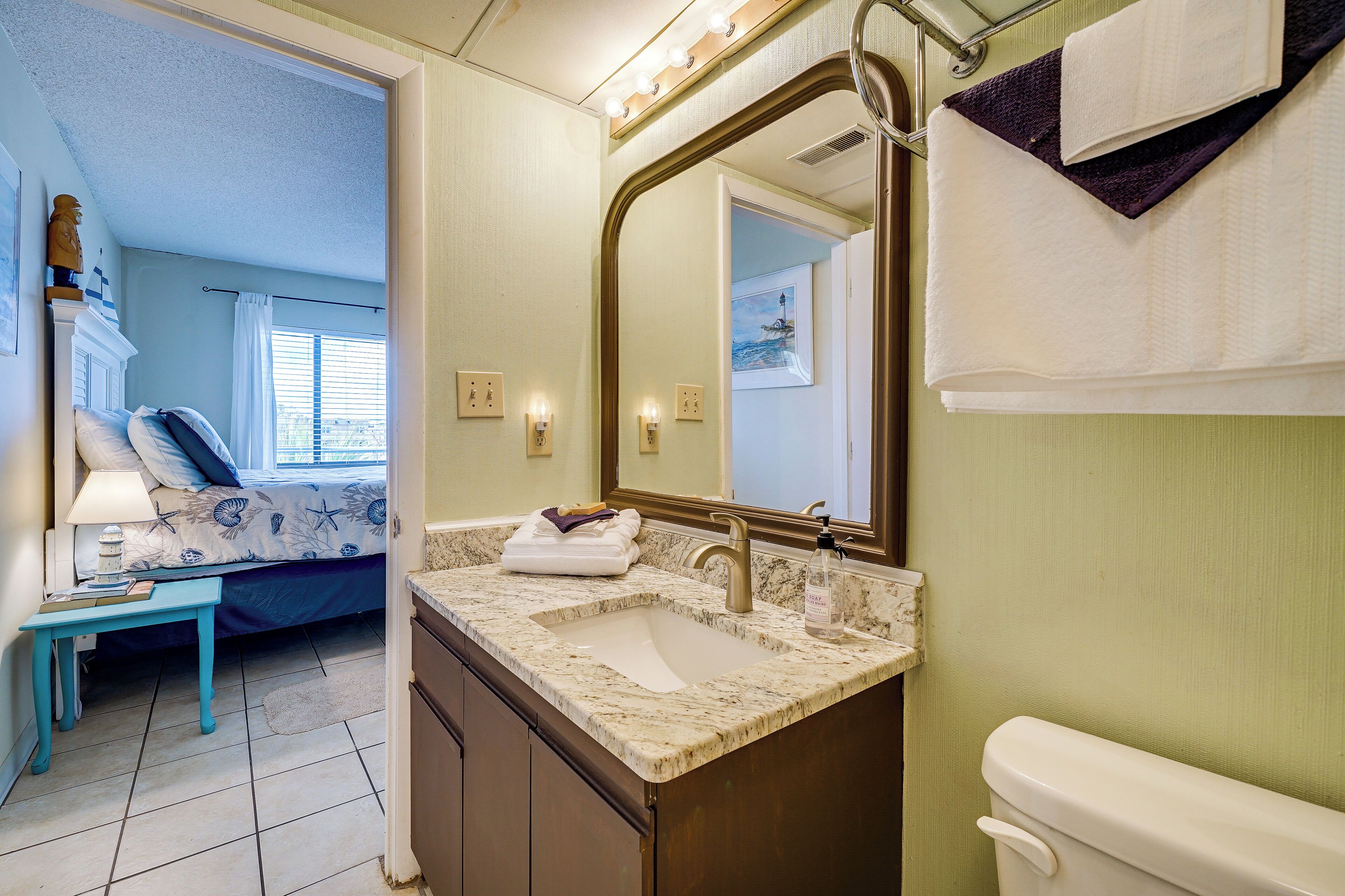 A bathroom with a sink, mirror, and toilet. There is a bed with a blue blanket and pillows in the adjacent room.